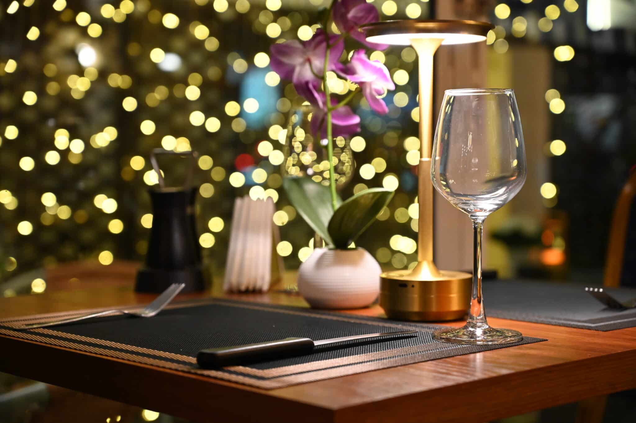 Candlelit restaurant table set for dinner with a wine glass and orchid against warm bokeh lights