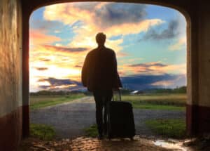 Traveller silhouetted with a wheeled suitcase walking towards a sunset sky at the end of a lane