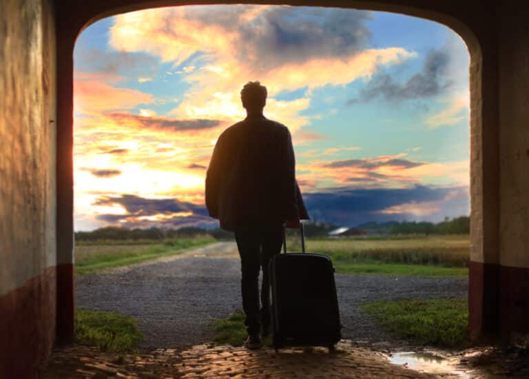 Traveller silhouetted with a wheeled suitcase walking towards a sunset sky at the end of a lane