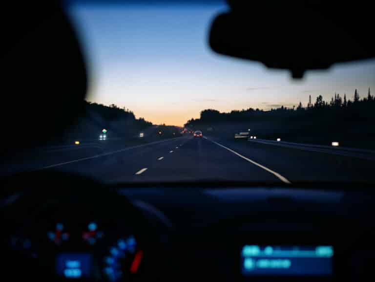 View through a car windscreen of a highway at dusk with tail lights ahead and a fading orange sky