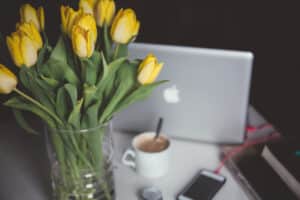 Laptop, coffee cup and notebook on a desk beside a vase of yellow tulips