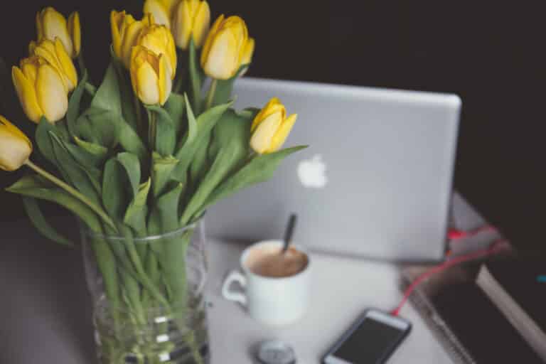 Laptop, coffee cup and notebook on a desk beside a vase of yellow tulips